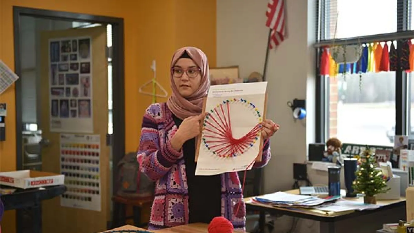 Noor Murteza stands in a colored knit sweater holding a pink fractal pattern, looking to side of camera