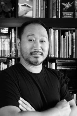Black and white photo of man standing wearing dark shirt standing in front of bookshelf