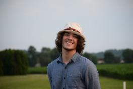 Braydon Tomak stands in field wearing button down shirt and hat, smiling at camera