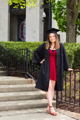 Kenzie Gelo stands on stairs smiling at camera, wearing graduation cap and gown