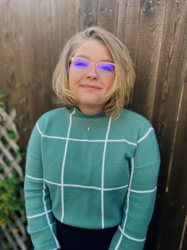 Megan Wanttie stands in front of wood fence wearing a green checkered shirt and glasses while looking at camera