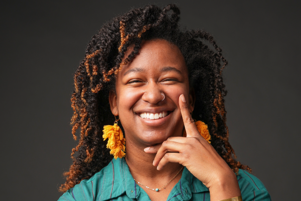 Woman in blue shirt with yellow dangling earrings, smiling at camera with hand on face