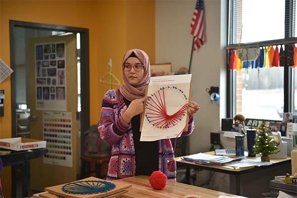 Noor Murteza stands in a colored knit sweater holding a pink fractal pattern, looking to side of camera