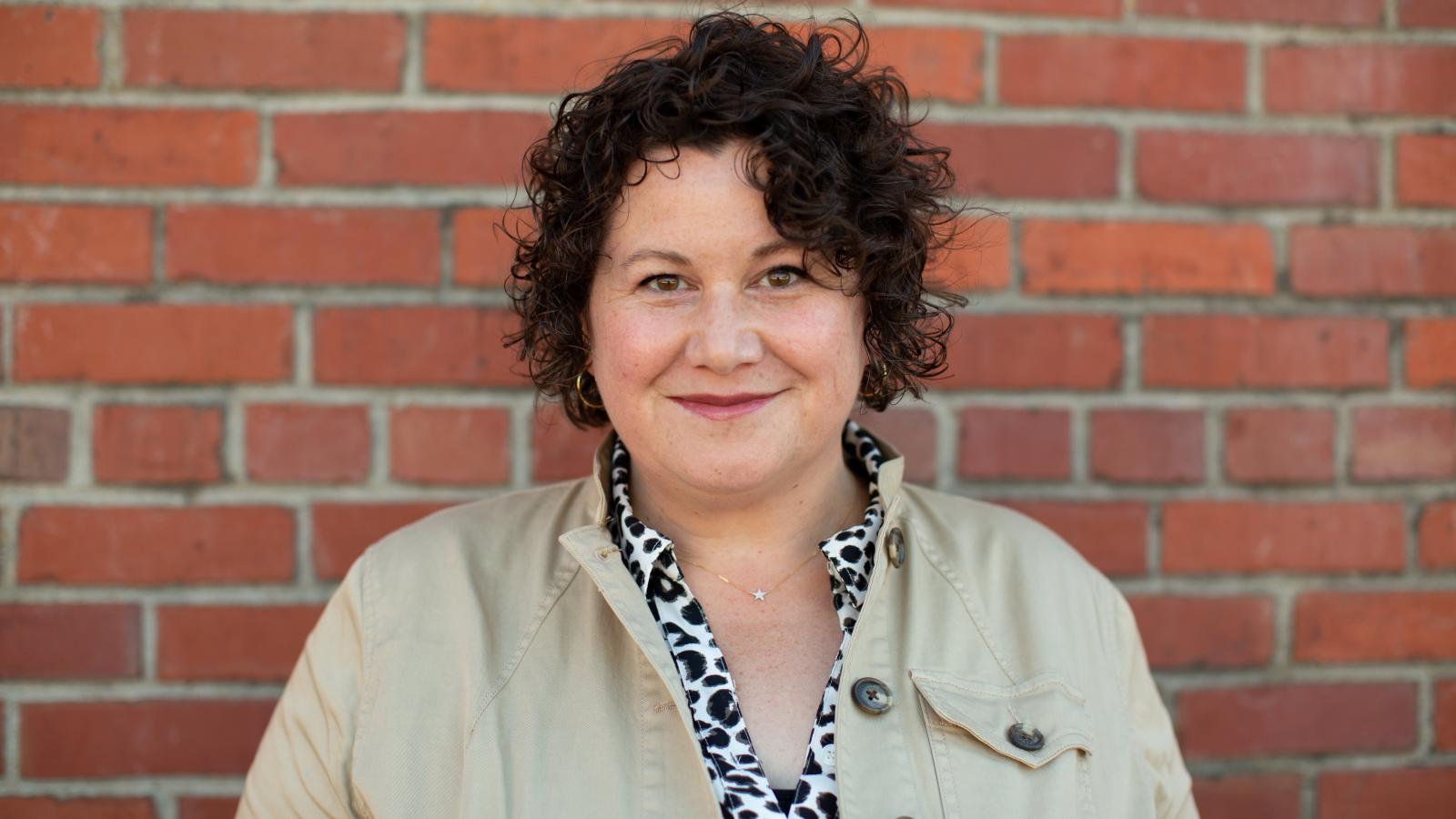 Woman with short, curly hair wearing khaki colored jackets smiles at camera while standing in front of brick wall